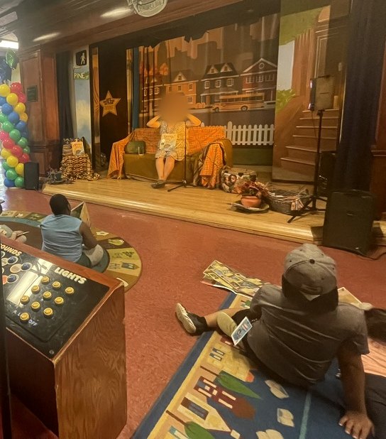 Story time on a theater stage with a balloon column and a neighborhood backdrop, performer's face blurred